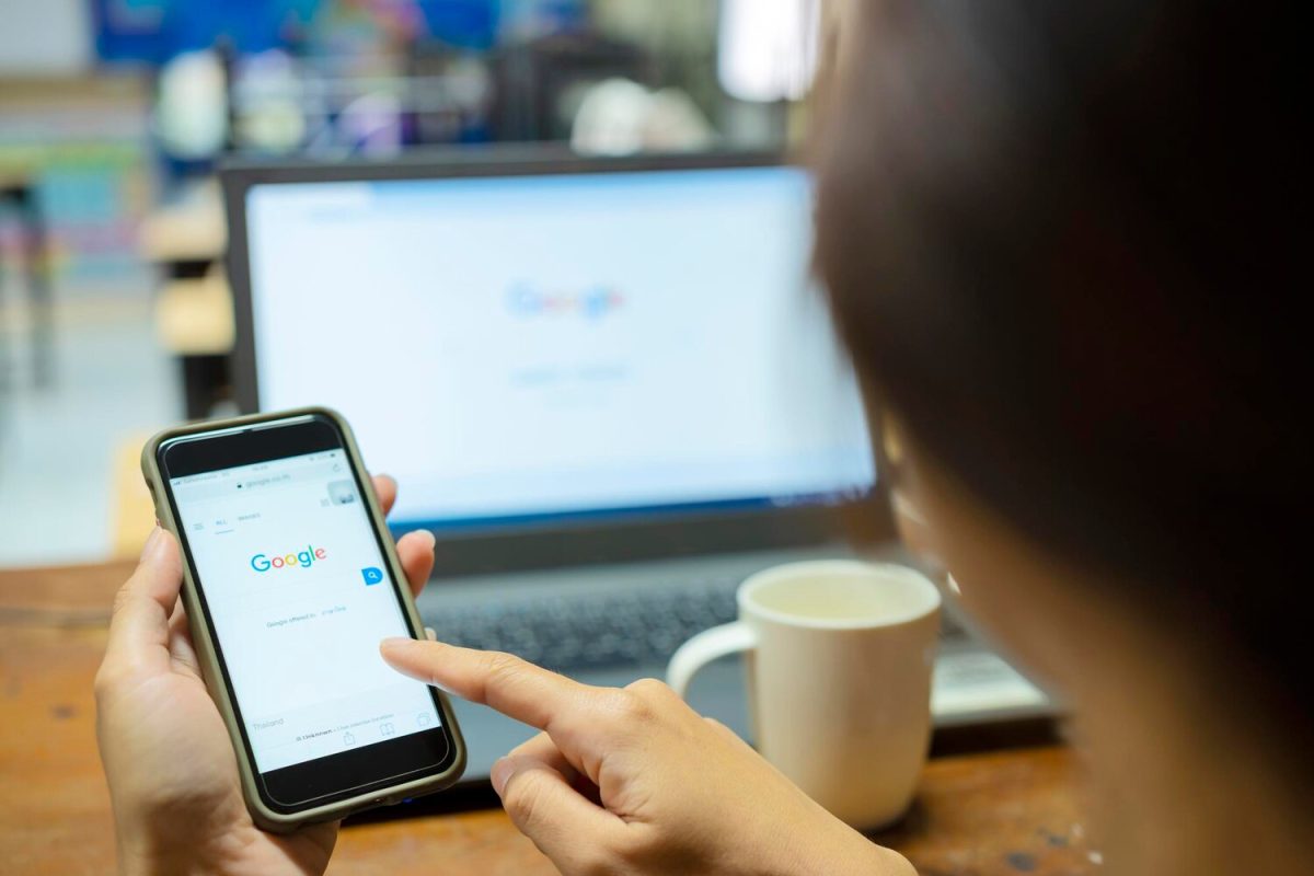 Egy személy okostelefonon görgeti a Google keresőoldalát, háttérben egy laptopon is látható a Google logó, SEO kulcsszavak keresése.