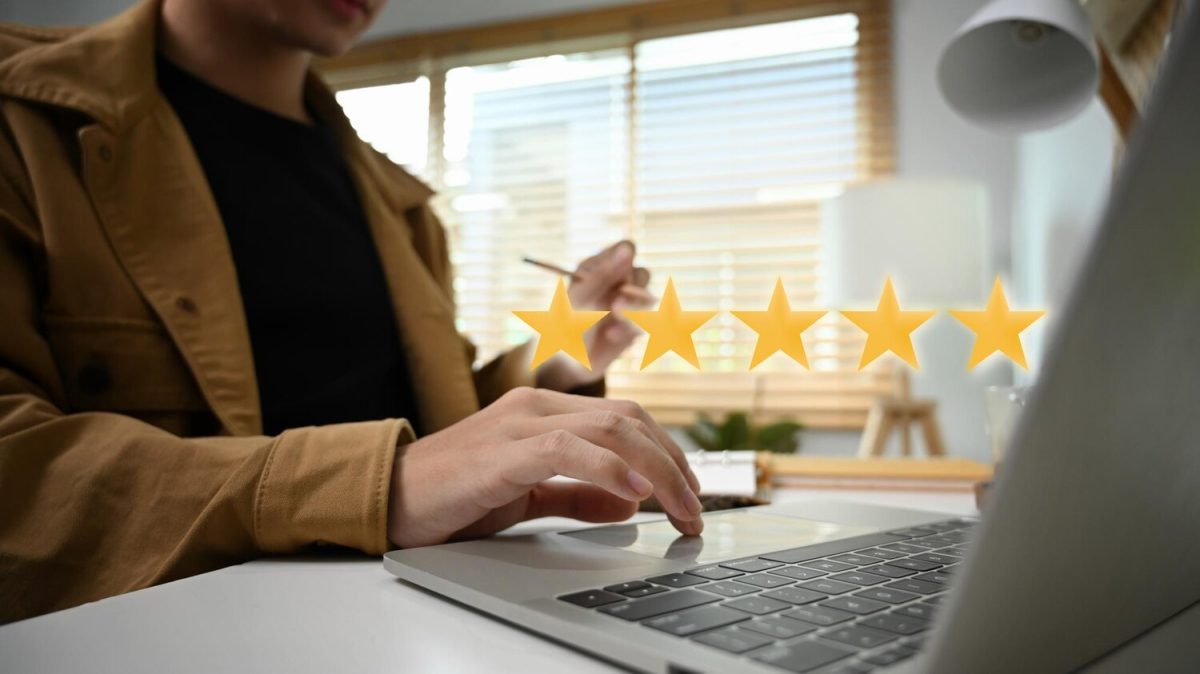 Egy személy laptopon gépel, miközben öt sárga csillag lebeg a képernyő felett, ami az online értékeléseket és a vevői visszajelzéseket szimbolizálja. A háttérben ablak és irodai környezet látható.