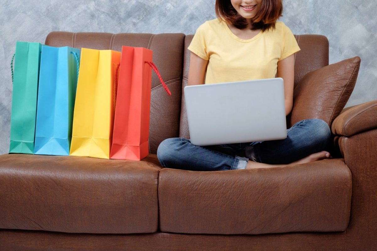 Fiatal nő laptoppal a kanapén, mellette színes bevásárlószatyrok online vásárláshoz.