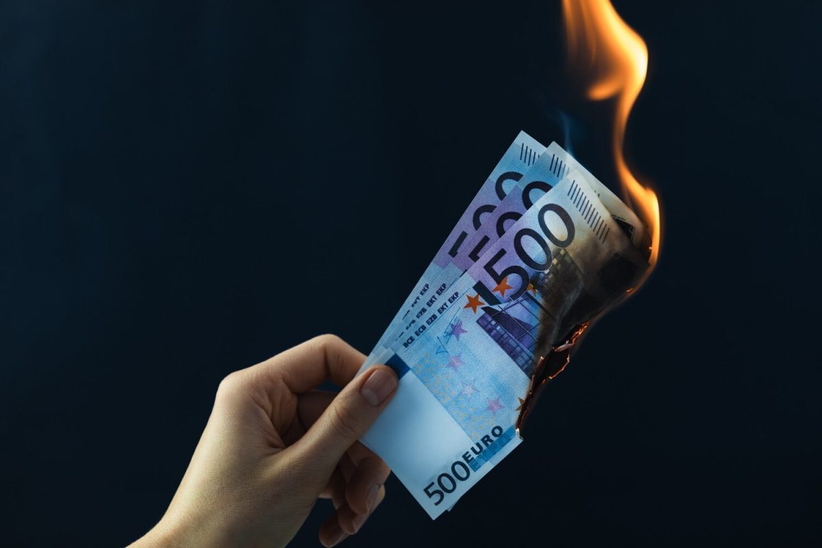 Egy kéz lángoló 500 eurós bankjegyeket tart sötét háttér előtt.