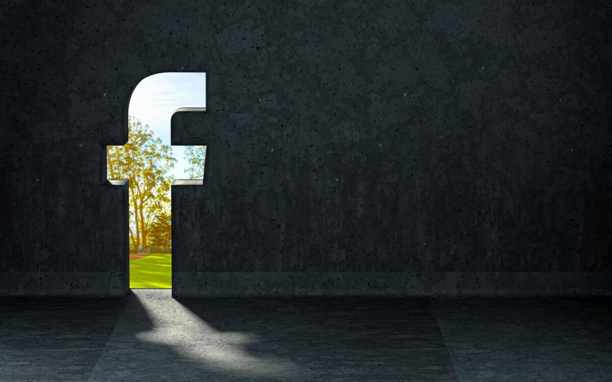 Sötét betonfal Facebook-logó alakú nyílással, amely napsütötte zöld természetre enged kilátást.