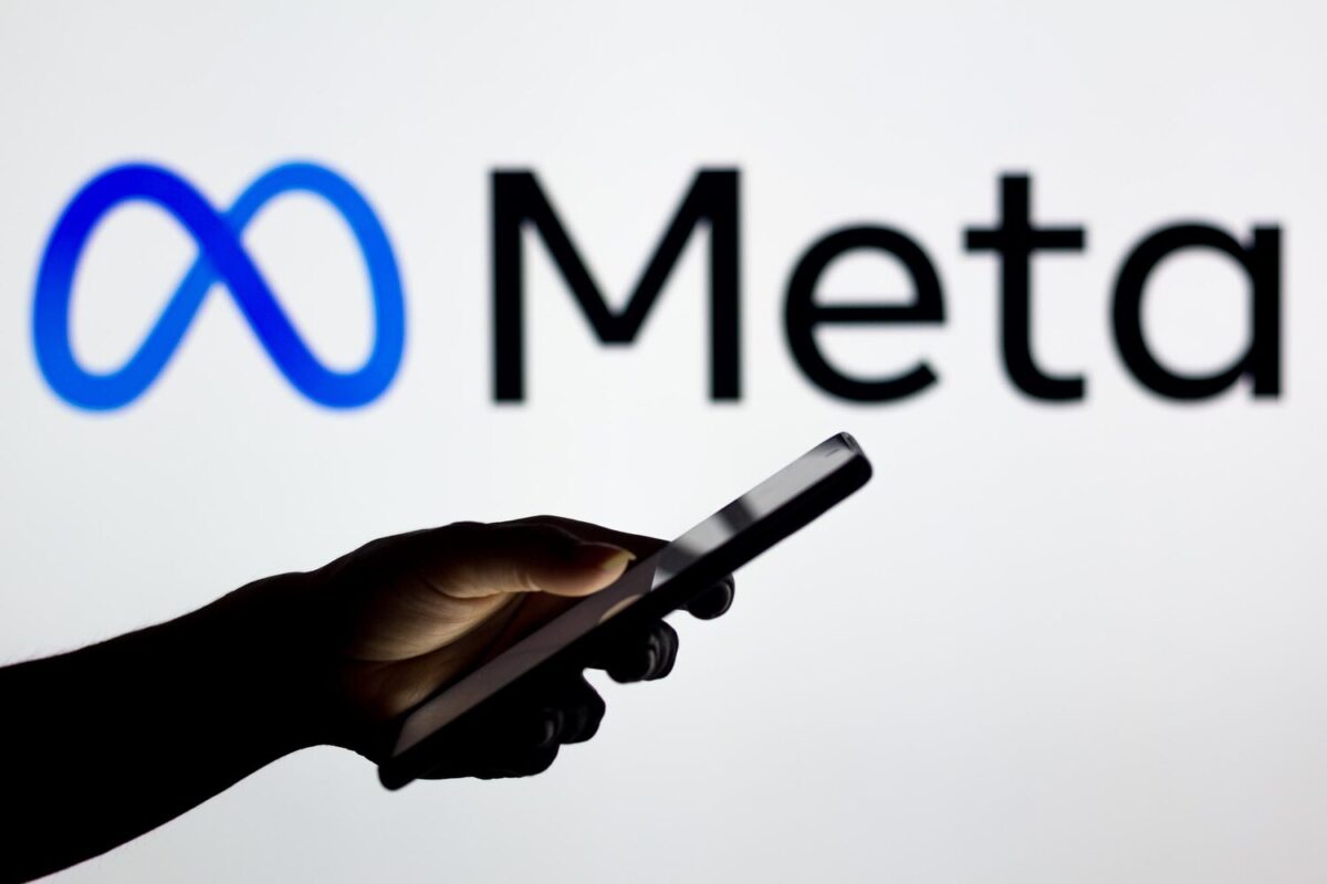 Okostelefont tartó kéz a háttérben megvilágított Meta logó előtt